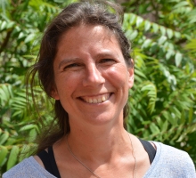 A headshot of Elly Moseley outside in the sunshine, in front of some foliage. She wears a black vest under a grey t-shirt, with a silver necklace, and smiles at the camera.