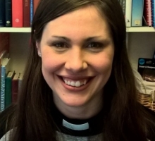 A headshot of Hannah Alderson, sitting in front of a bookcase of text books. She wears a dog collar and smiles at the camera.
