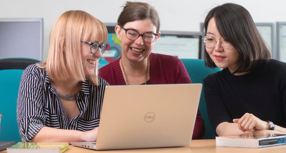 Three women sat around a laptop, smiling. They have a book next to them.